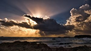 Stunning sunset casting sunrays through clouds over ocean with rocky shore.