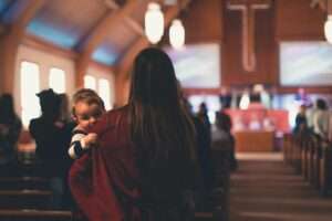 Woman carrying a child inside a church with people worshipping in the background.