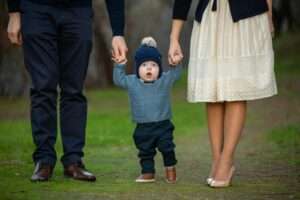 A cute toddler holds hands with parents while walking in a park, wearing a beanie.