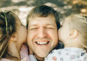 A joyful father shares a loving moment with his daughters as they kiss his cheeks.