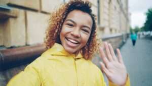 Joyful woman waving at camera outdoors in yellow raincoat. Bright happy moment captured.
